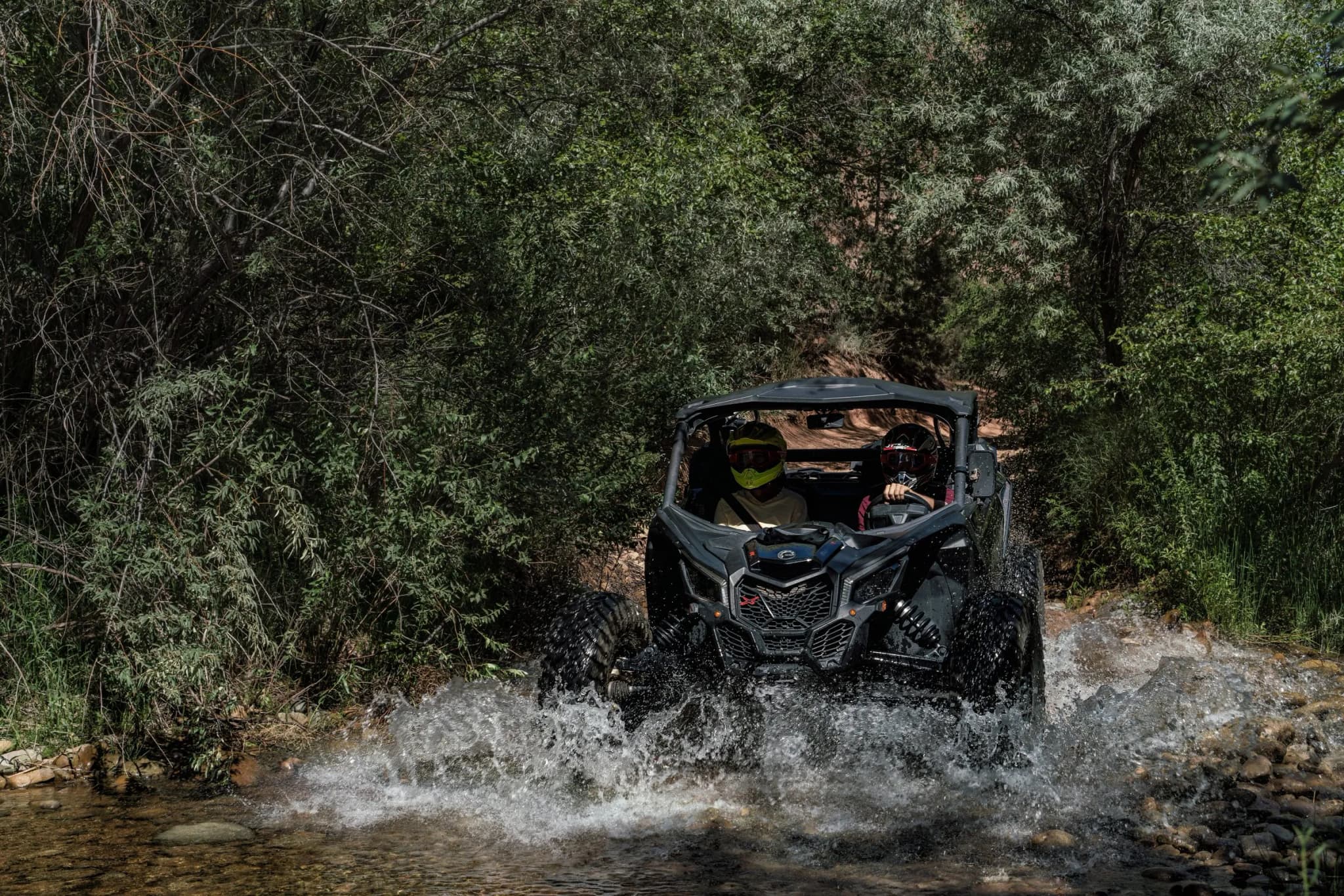 Two helmeted riders splash a Can-Am X3 through a shallow creek lined with trees