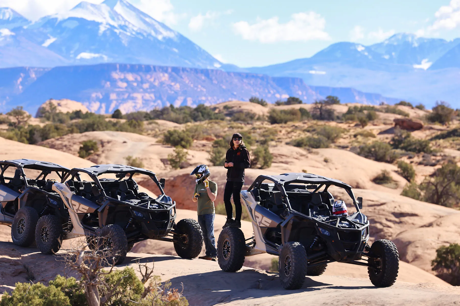 Riders beside parked Can-Am X3 side-by-sides on slickrock, with snow-capped mountains in the distance