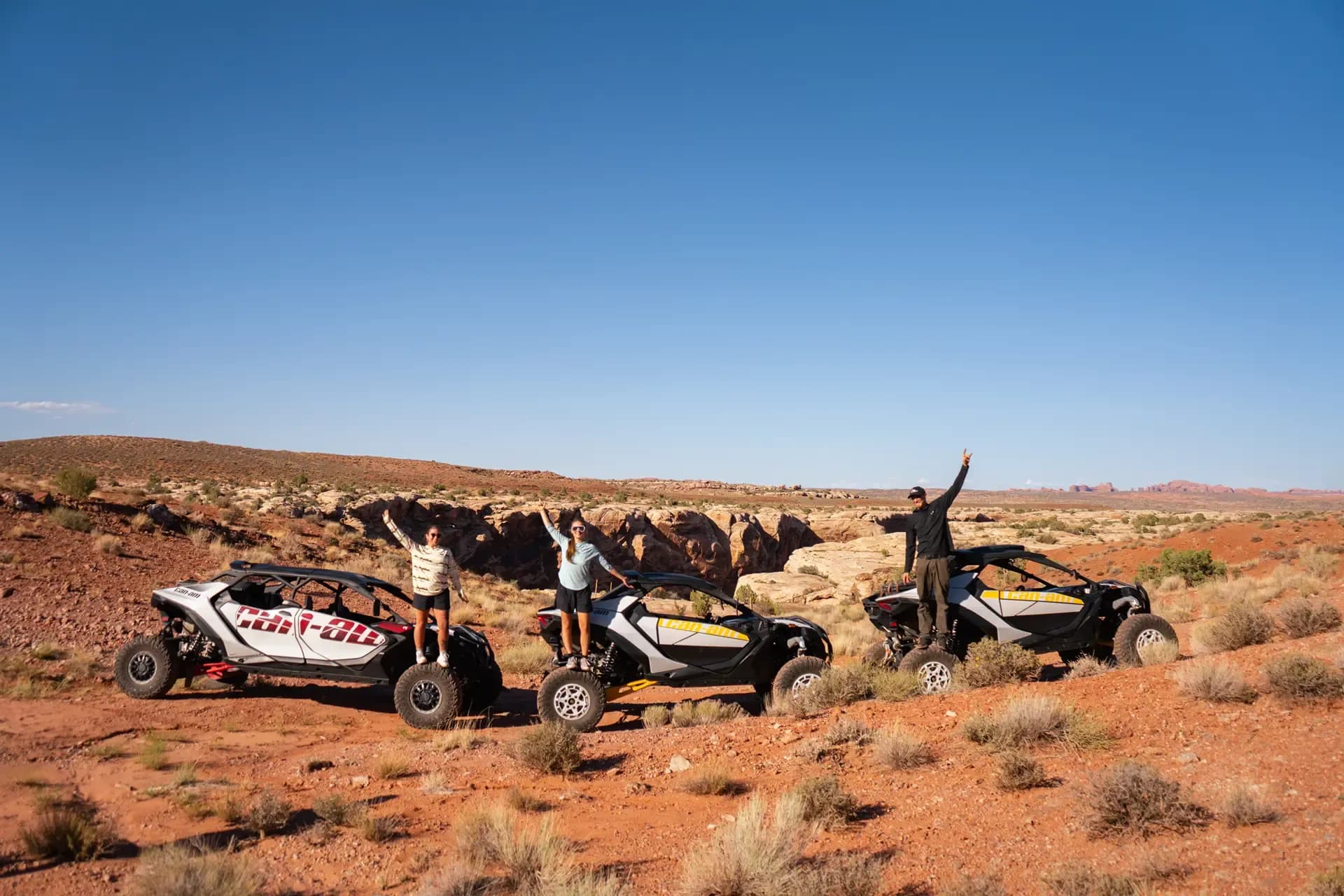 Three guests cheering on top of parked Can-Am UTVs on a red-rock trail
