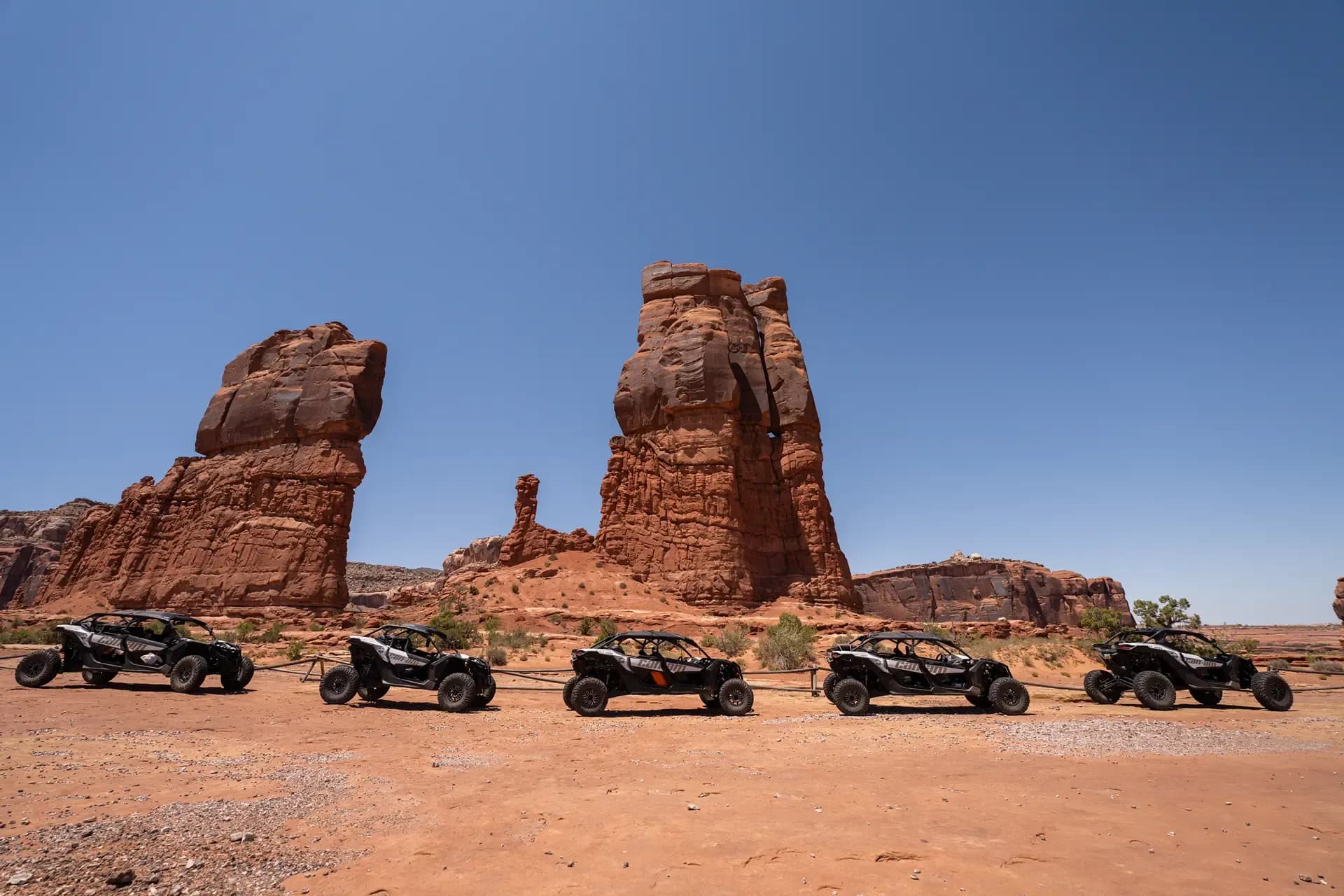 Five black Can-Am UTVs parked in a row before twin red rock sandstone towers