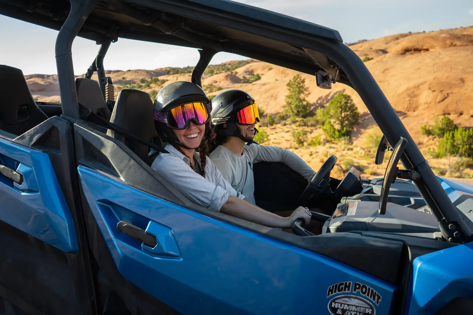 Two guests smiling in helmets and goggles riding in a blue Can-Am UTV with desert hills behind
